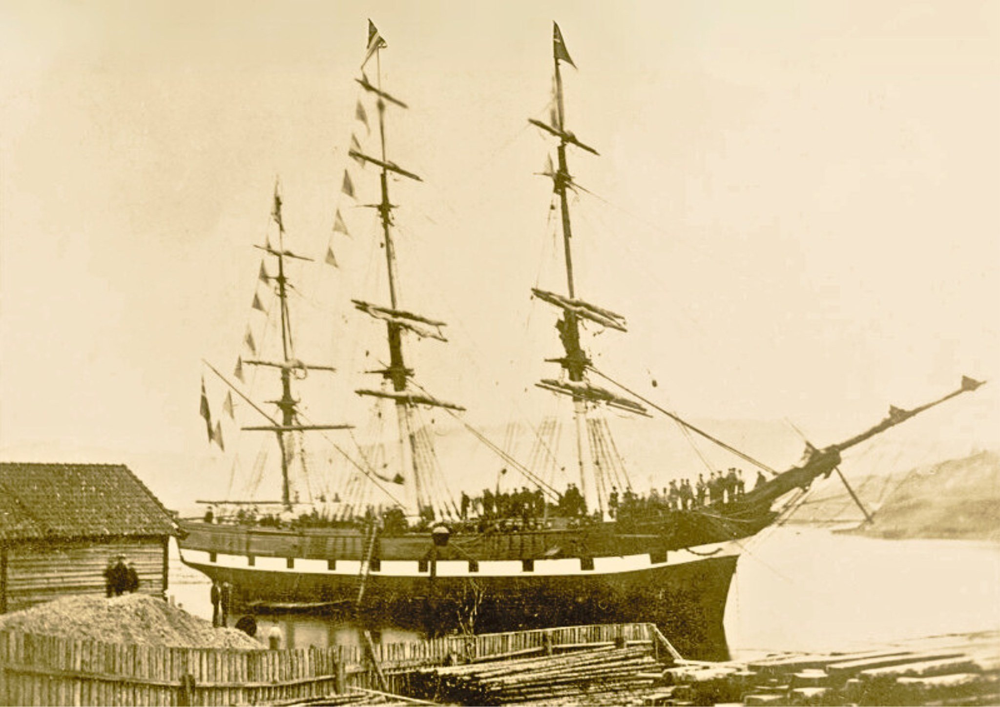 Gammelt seilskip ved kai, med mange seil og flere personer ombord, historisk sjøfartsmotiv.