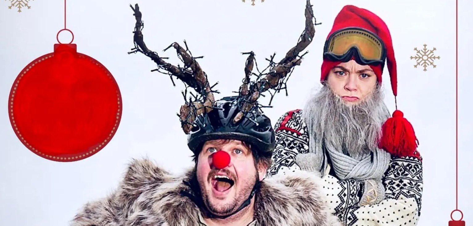 To personer utkledd som reinsdyr og nisse, med julepynt i bakgrunnen. Festlig og humoristisk juletema.