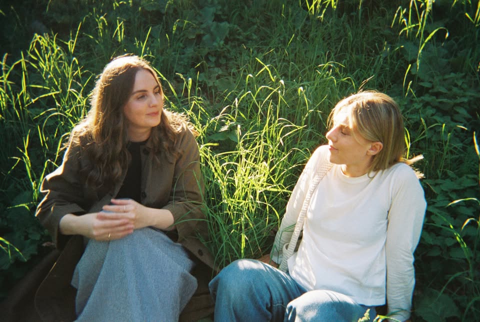 To kvinner sitter i solfylt gress, kledd i avslappet stil, og prater i en naturlig setting.