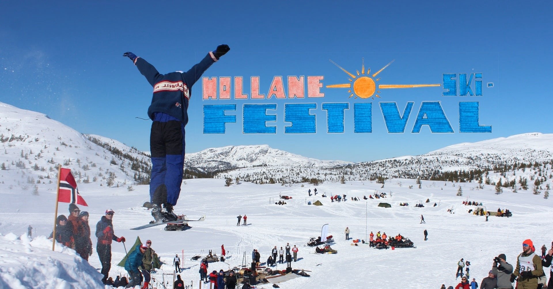 Snøbrettkjører svever under klar blå himmel på Hollane Ski Festival. Snødekte fjell i bakgrunnen.