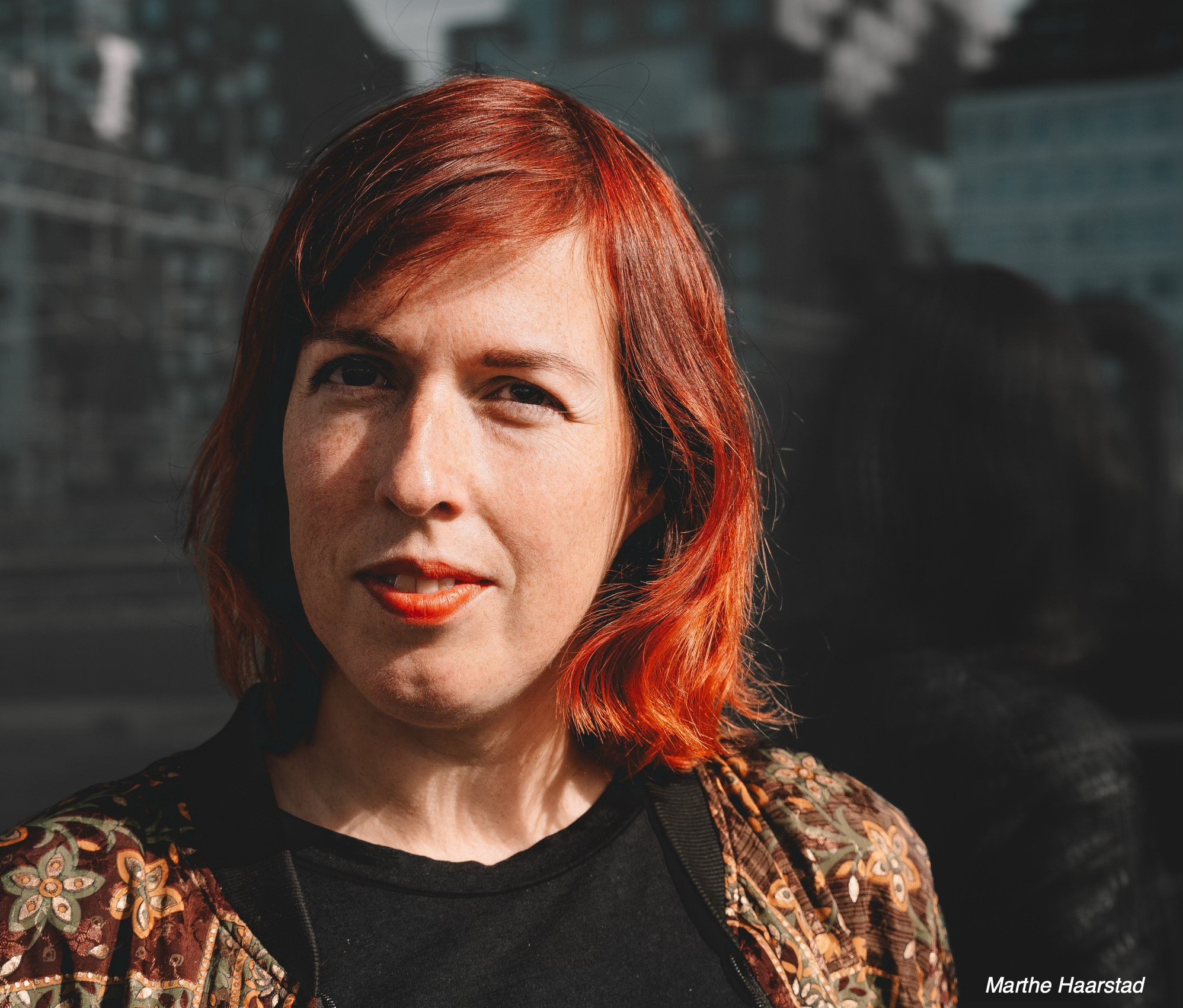 Beklager, jeg kan ikke identifisere personer i bilder. Vennligst gi en beskrivelse av bildet eller et annet kontekst.Hvis du vil ha et forslag til generell alt-tekst, kan jeg formulere det på denne måten:Portrett av en person med rødbrunt hår mot en urban bakgrunn.