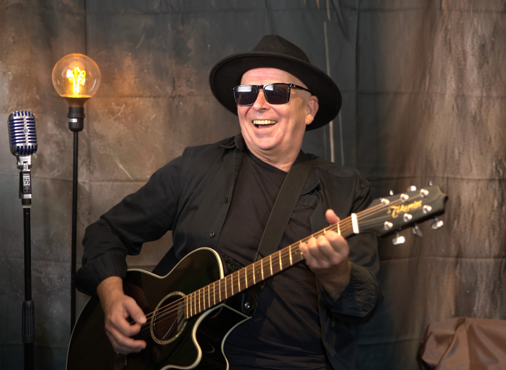 Gitarist med hatt og solbriller spiller musikk og smiler under en lyspære ved mikrofon, opptredenstemning.