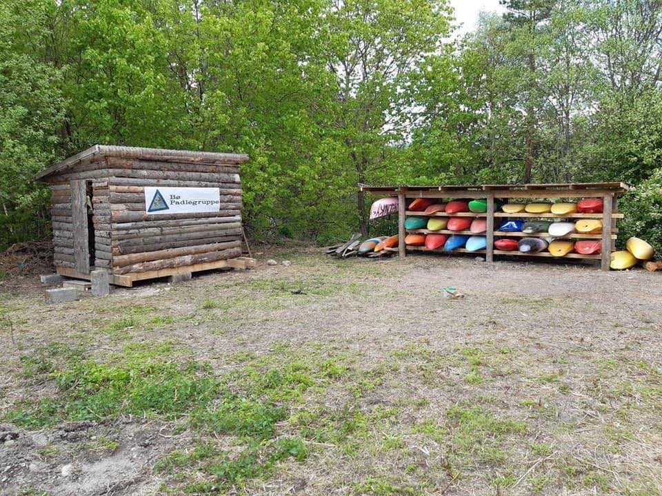 Uthus med kajakker i farger fra Bø Padlegruppes utleie, omgitt av grønn skog.