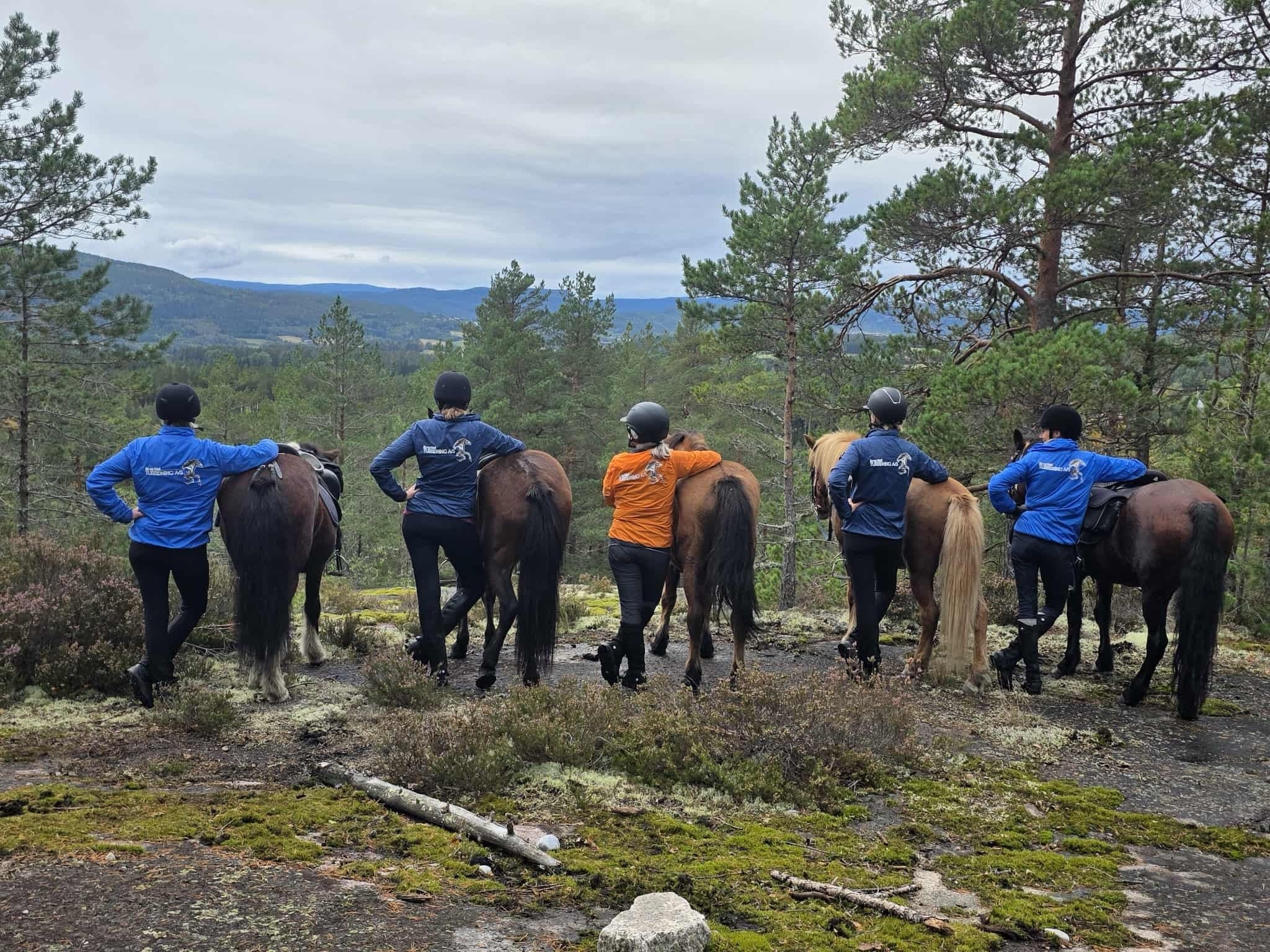 Ryttere står med hester i norsk skog, iført fargerike jakker, ser utover vakker natur.