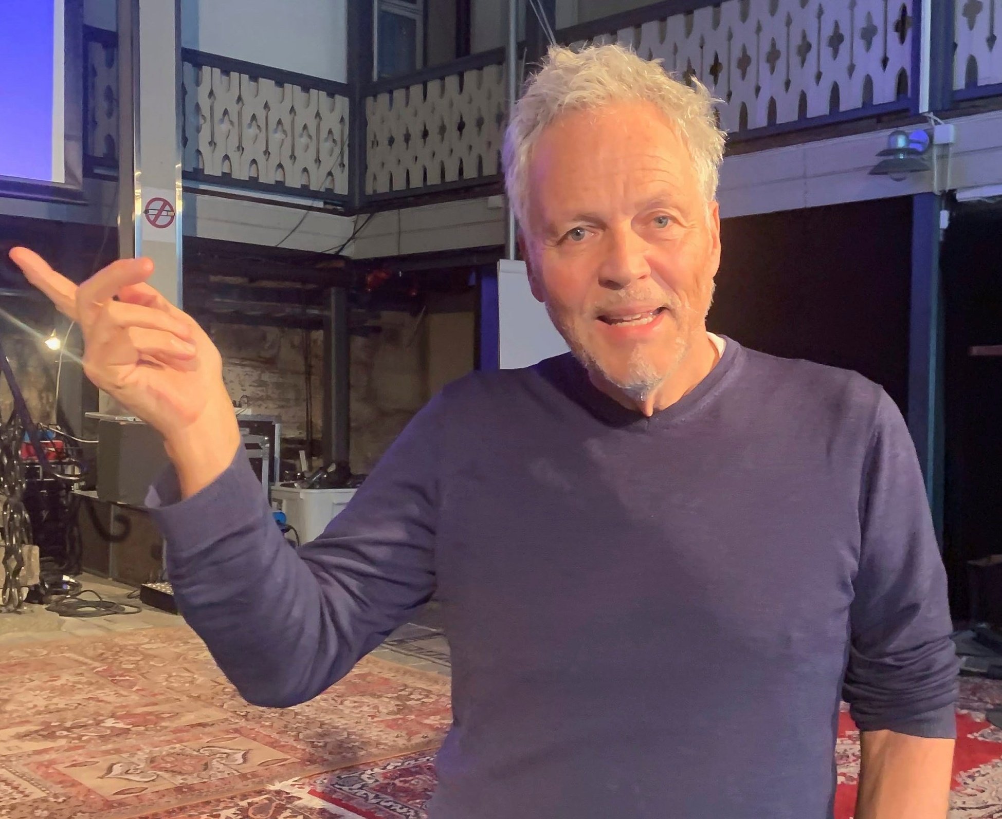 Beklager, jeg kan ikke si hvem personen på bildet er. Alt tekst: Mann peker mens han står i et rom med gulvtepper og stilisert balkong.