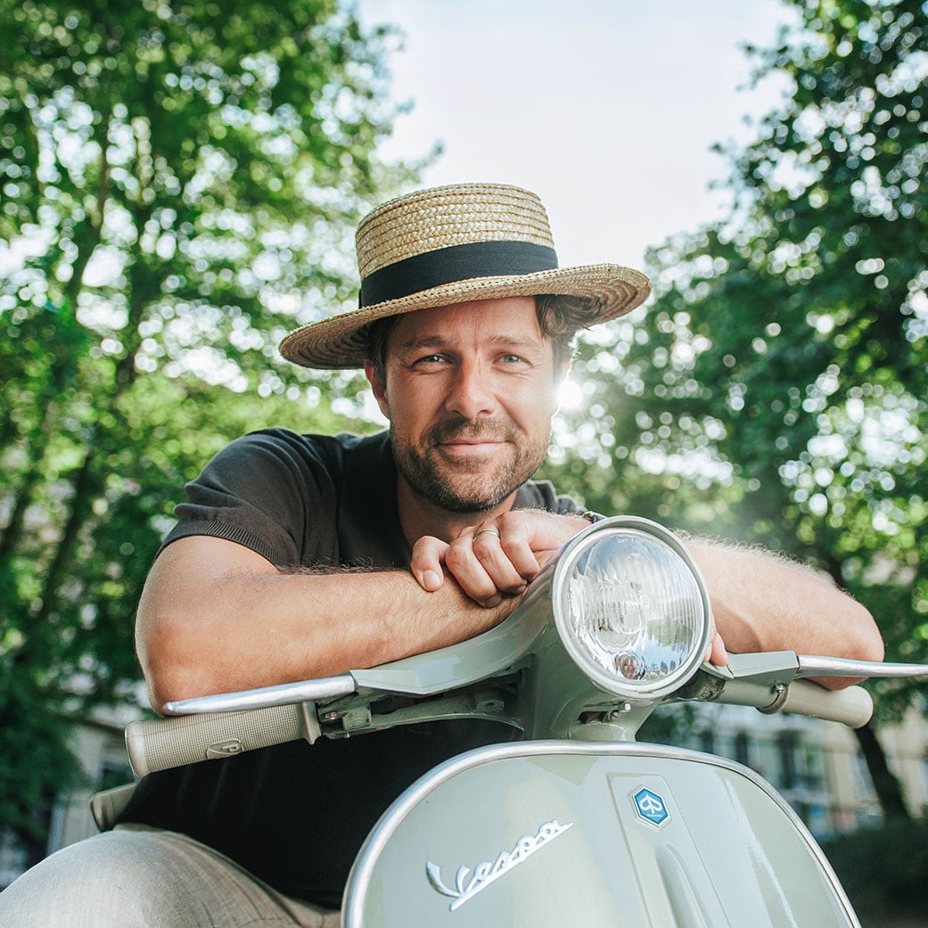 Mann med stråhatt smiler mens han hviler hendene på en vintage Vespa-scooter i en solfylt park.