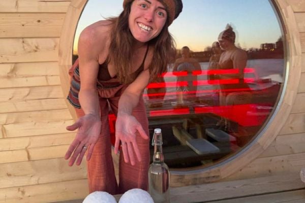 Ung kvinne smiler og viser frem snøballer og en flaske, med en rød bakgrunn av saunaen.