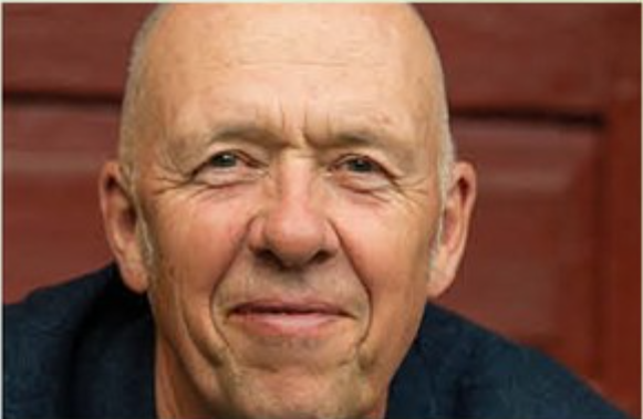 Portrett av en smilende middelaldrende mann med kort hår, iført mørkeblå jakke og bakgrunn i tre.