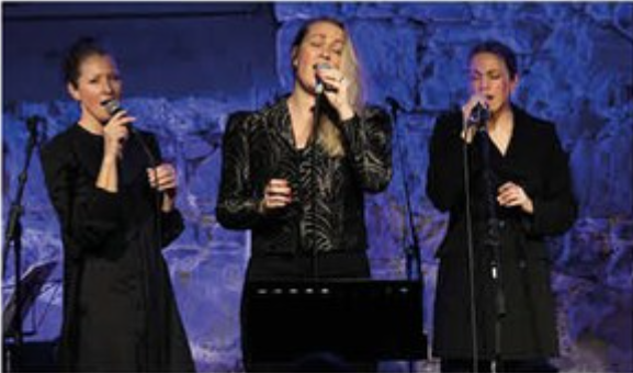 Tre kvinner synger på en scene, kledd i svart, med blåaktig lys i bakgrunnen. Livlig musikkopplevelse.