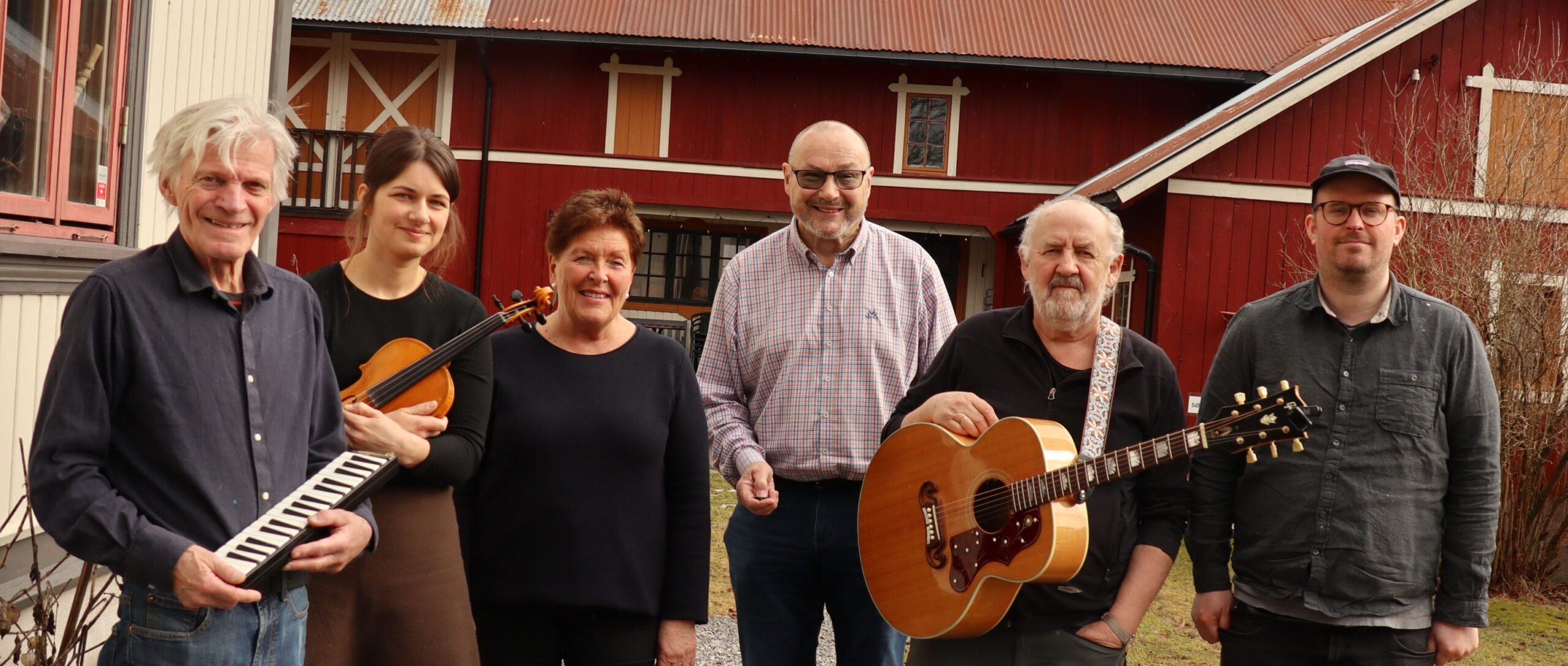 En gruppe musikere poserer foran en rød gård, med instrumenter som fiolin og gitar.