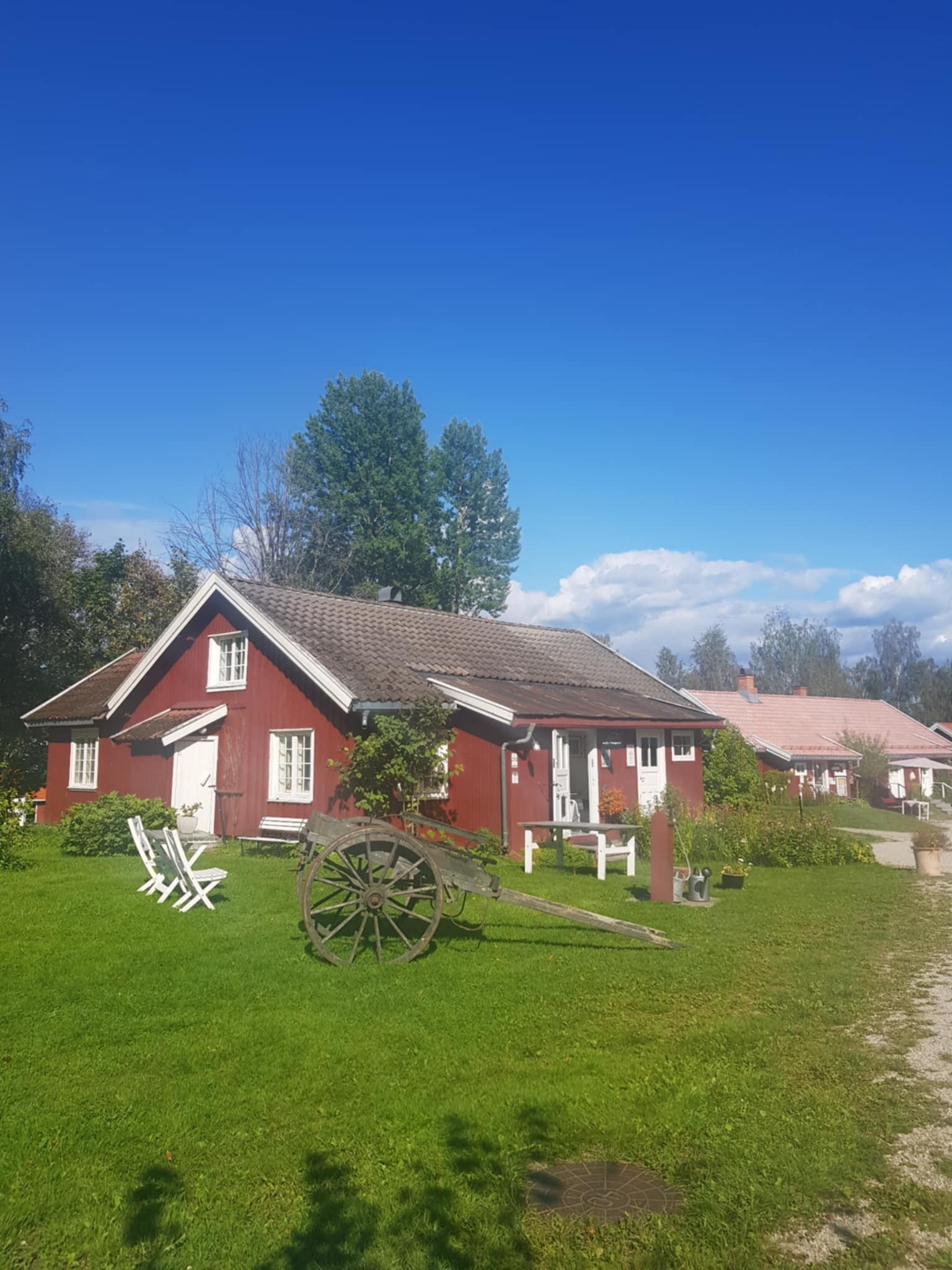 Rødt hus med hage, gamle redskaper og blå himmel, omkranset av frodige trær og grønt gress.