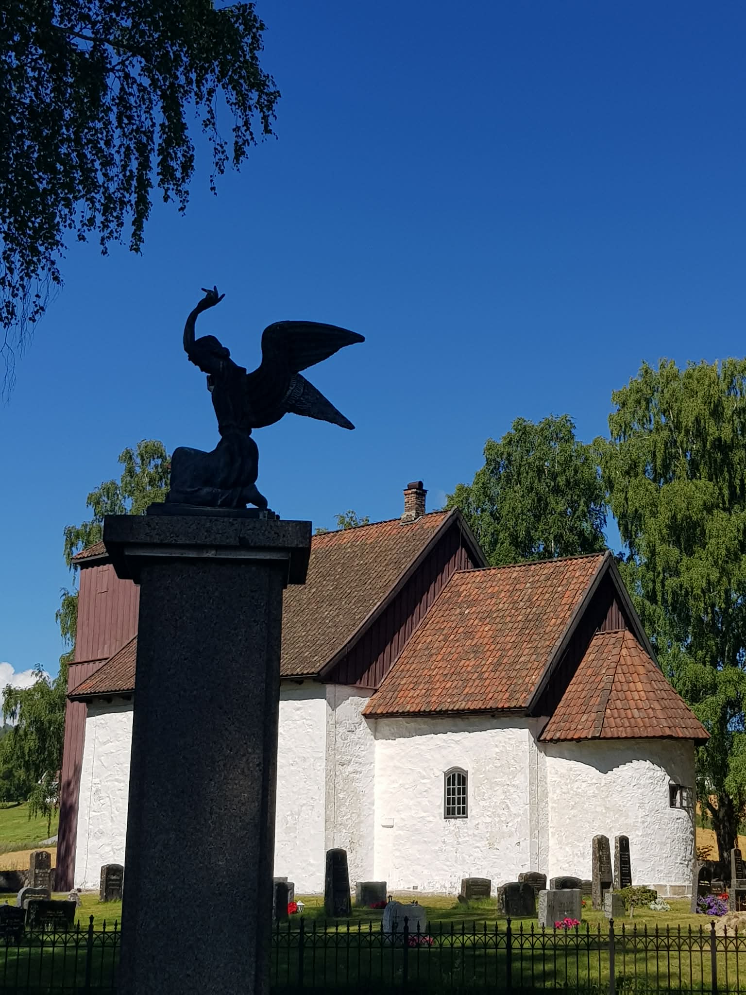 Bilde av en engelstatue foran en gammel kirke med gravsteiner og grønne trær under en klar blå himmel.