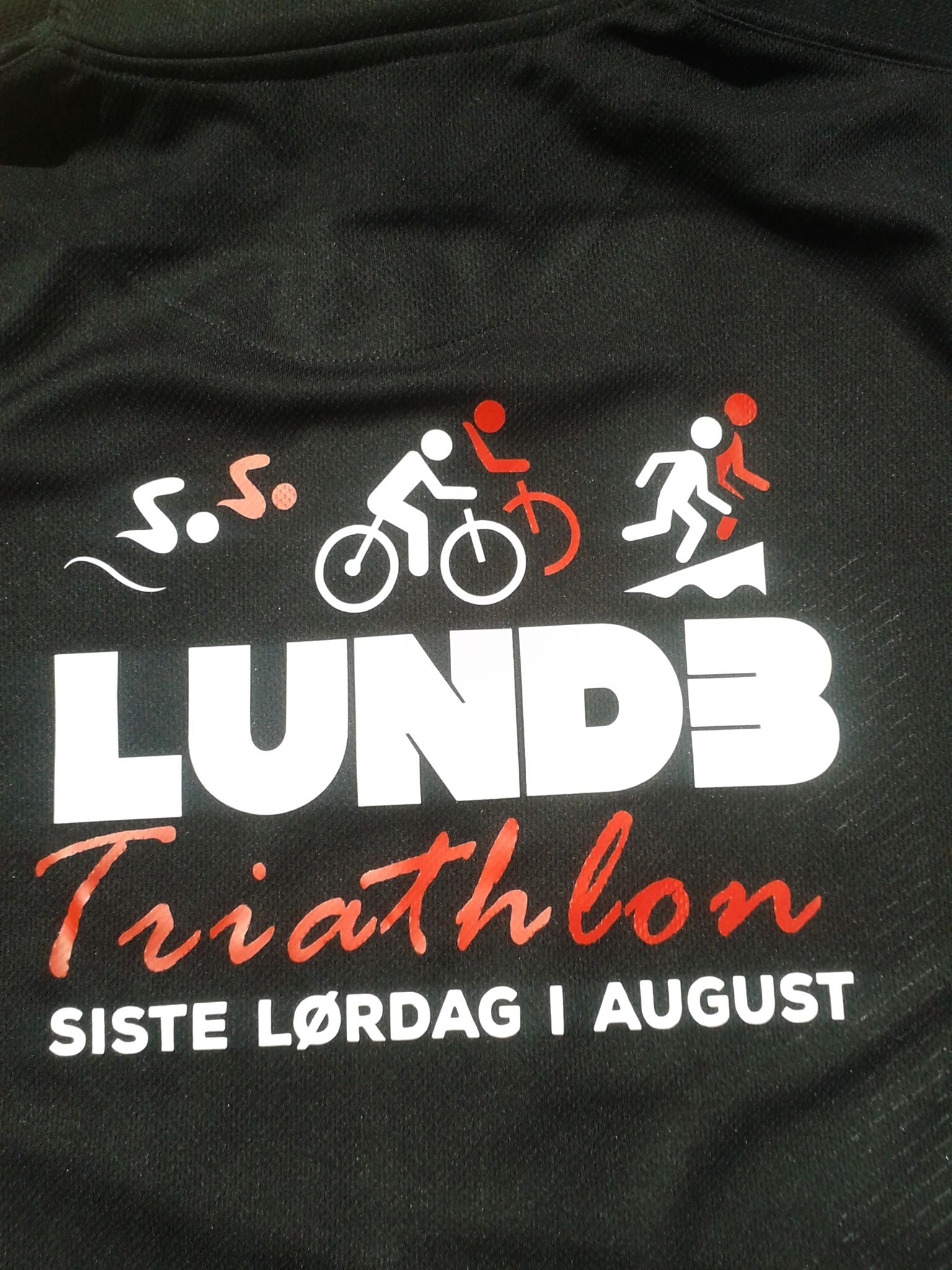 Alt-tekst: LUND3 Triathlon på en svart t-skjorte med dato: siste lørdag i august, med symboler for svømming, sykling og løping.