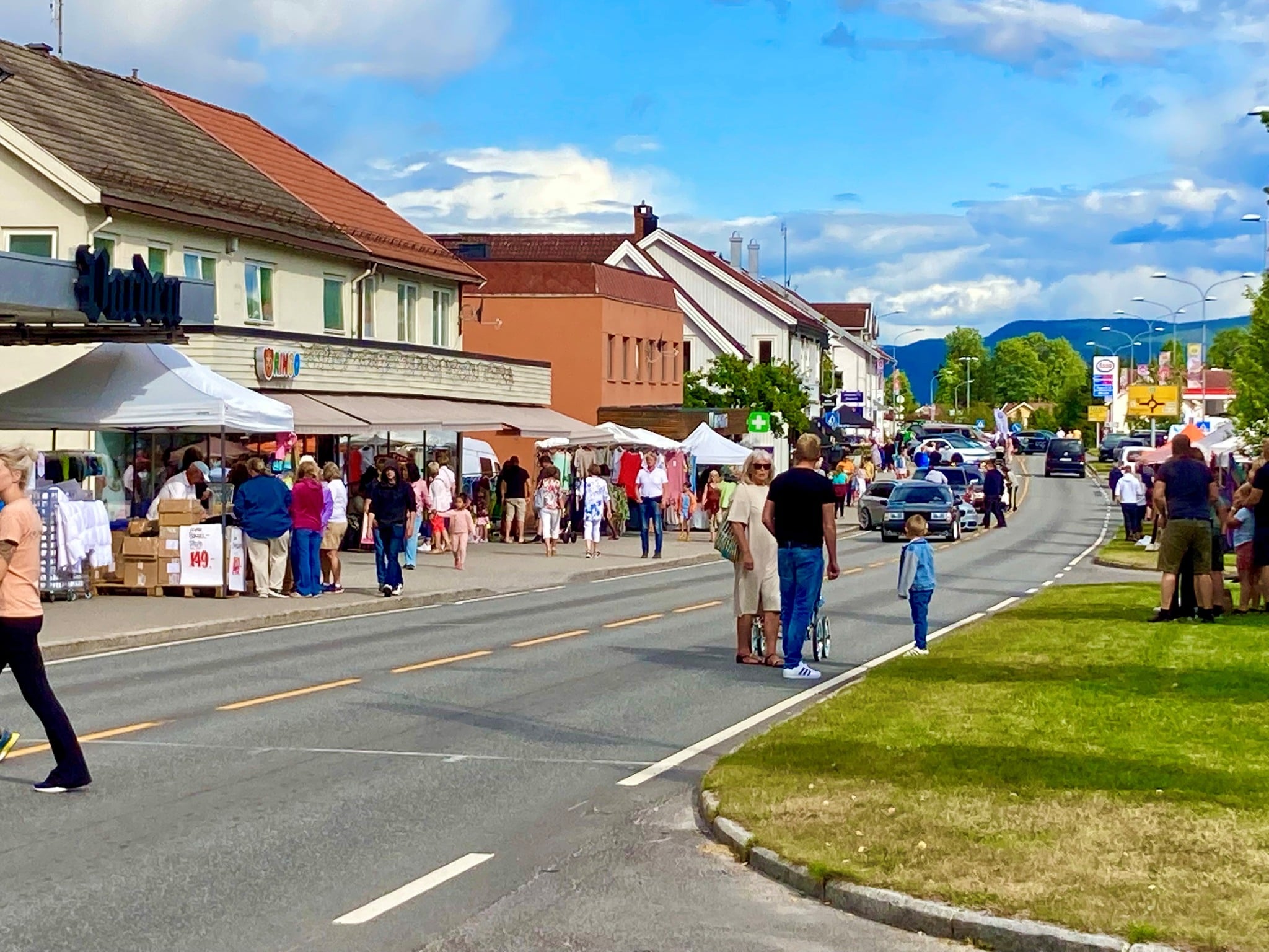Bilde av en livlig gate i Norge med mennesker, boder og handleturer på en solfylt dag.