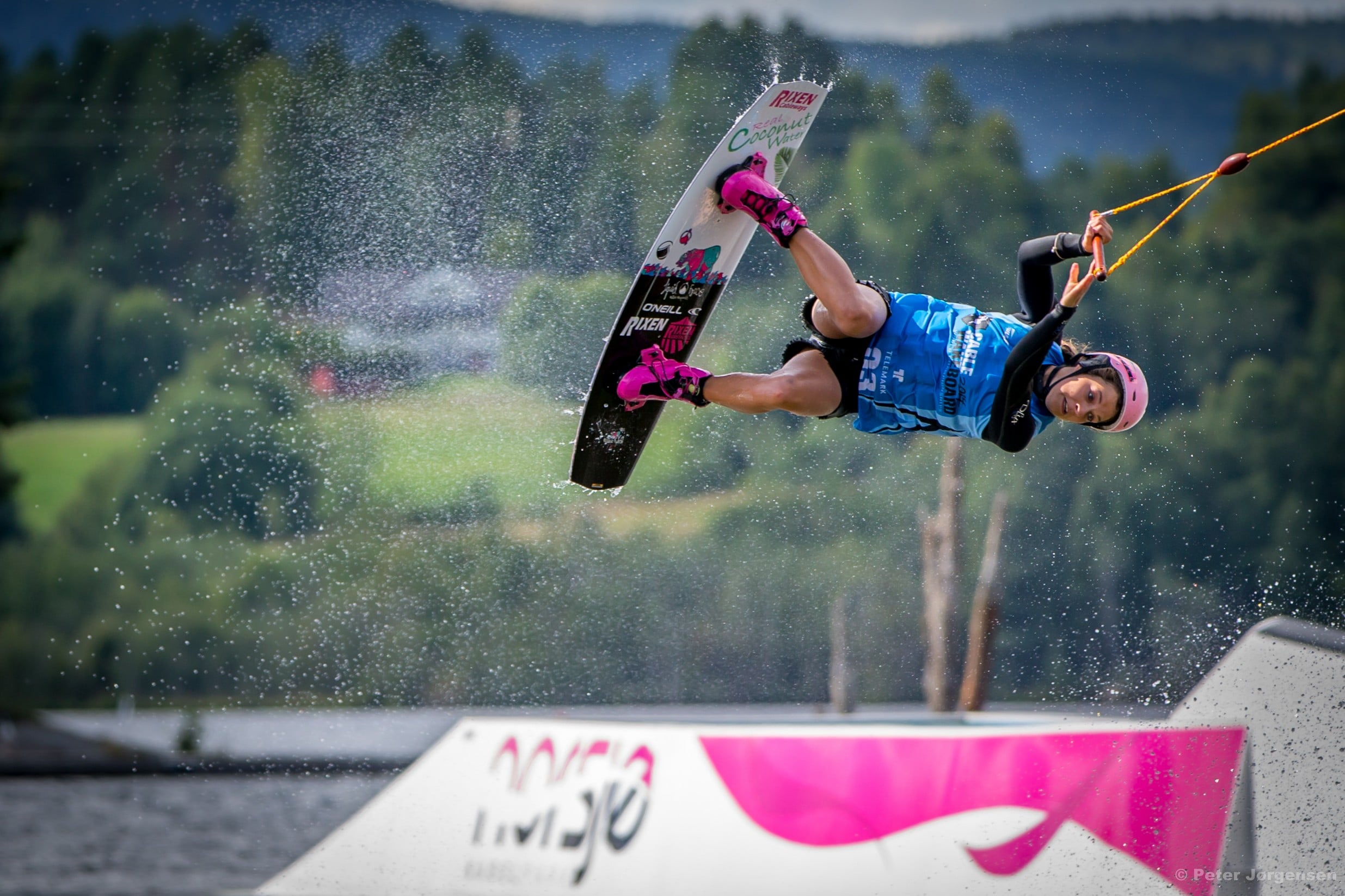 Kvinne utfører et spektakulært wakeboardtrick over vannet, omringet av sprut og grønne omgivelser.