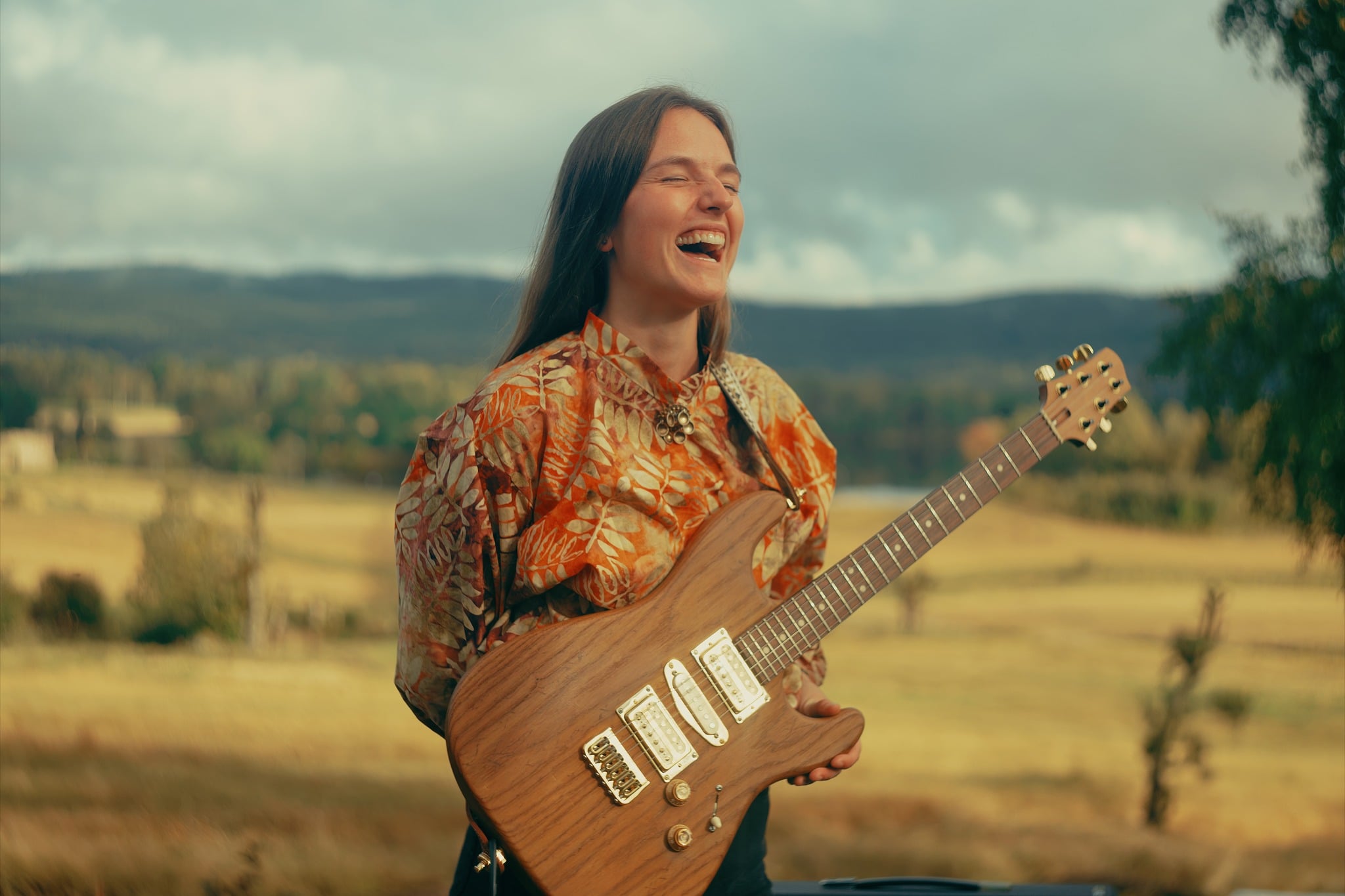 Ung kvinne med gitar, smilende i et vakkert landskap, iført fargerik skjorte. Musikk og glede i naturen.