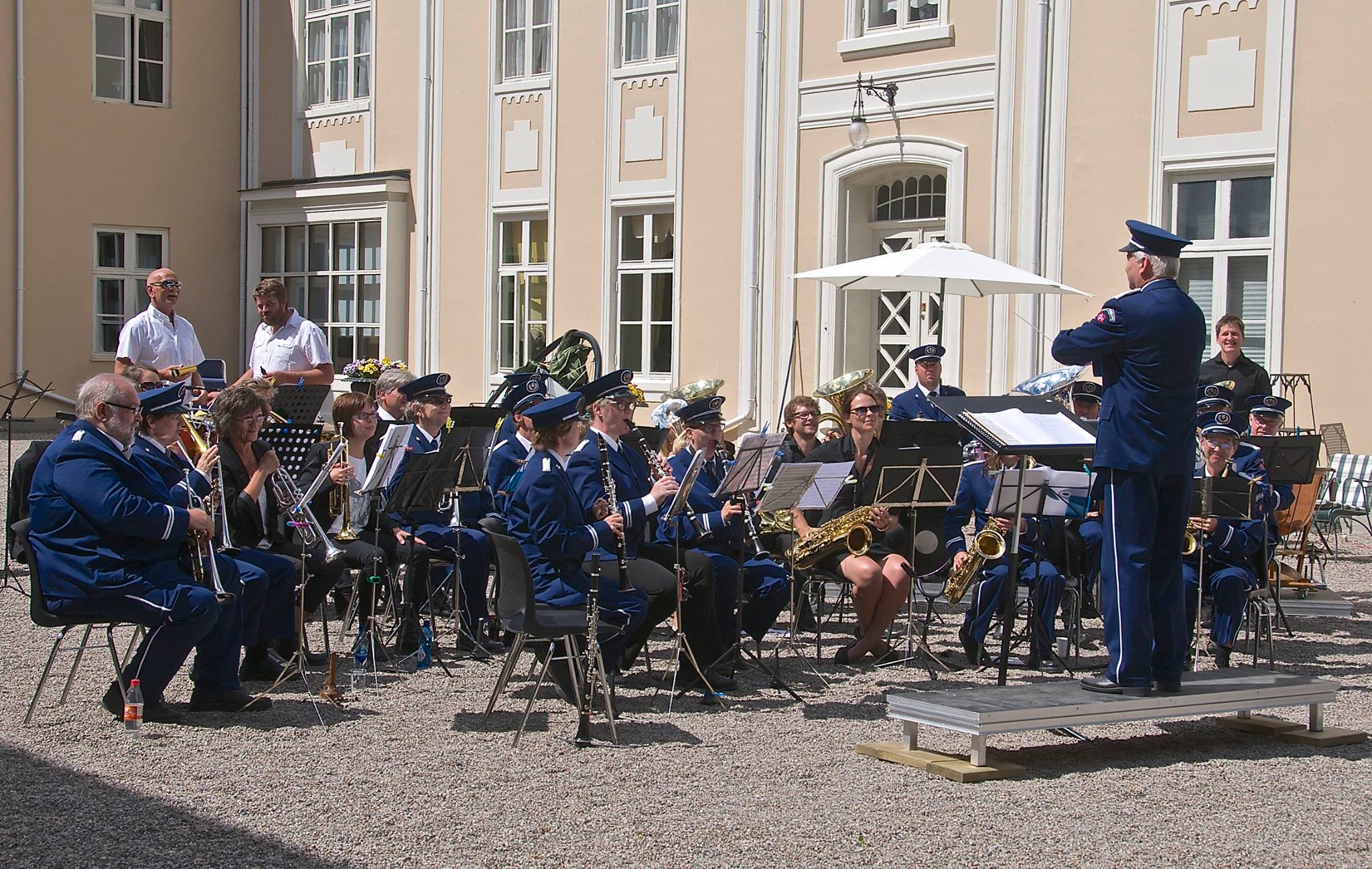 Musikere i uniform spiller i solfylt bakgård, dirigert av en mann foran orkesteret.