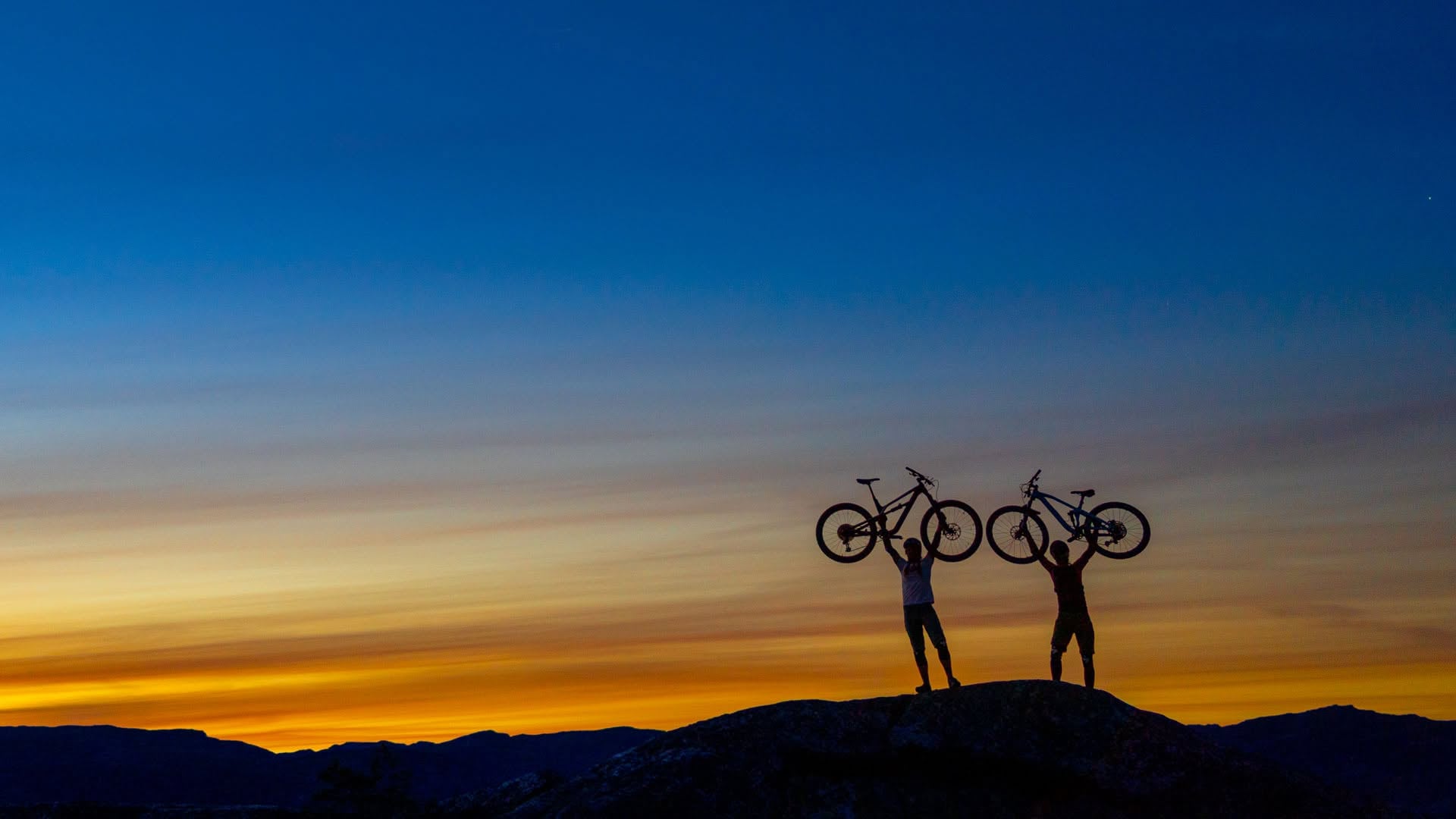 To personer på en høyde som hever syklene sine mot en vakker solnedgang i fjellene.
