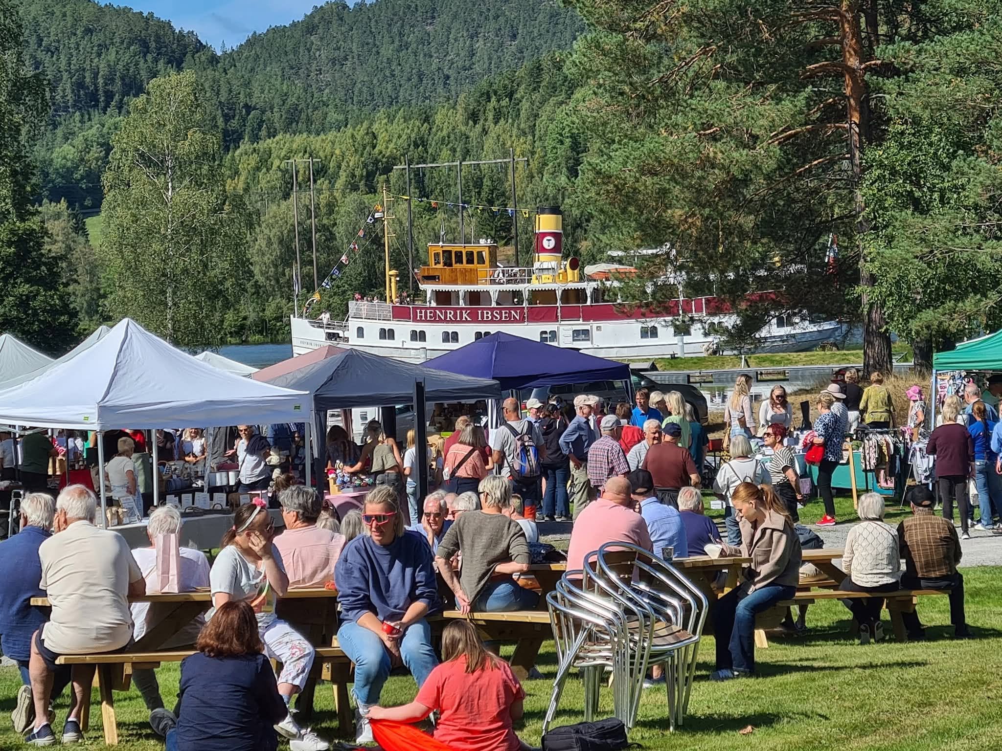 Markedsdag ved vannet med folk som nyter solen, boder og dampbåten Henrik Ibsen i bakgrunnen.