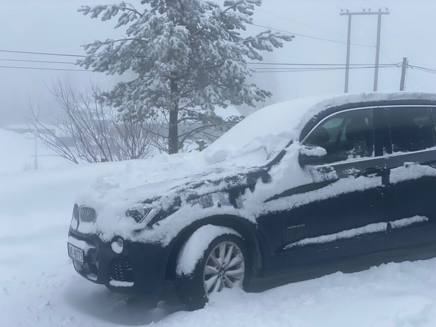 Bilde av en svart bil dekket av snø i en vinterlig landskap, med trær og strømkabler i bakgrunnen.